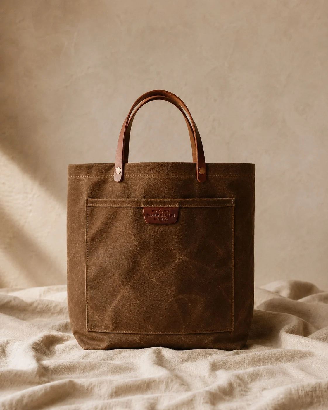 The Field Tan Coal Tote standing upright on linen against a warm plaster wall.