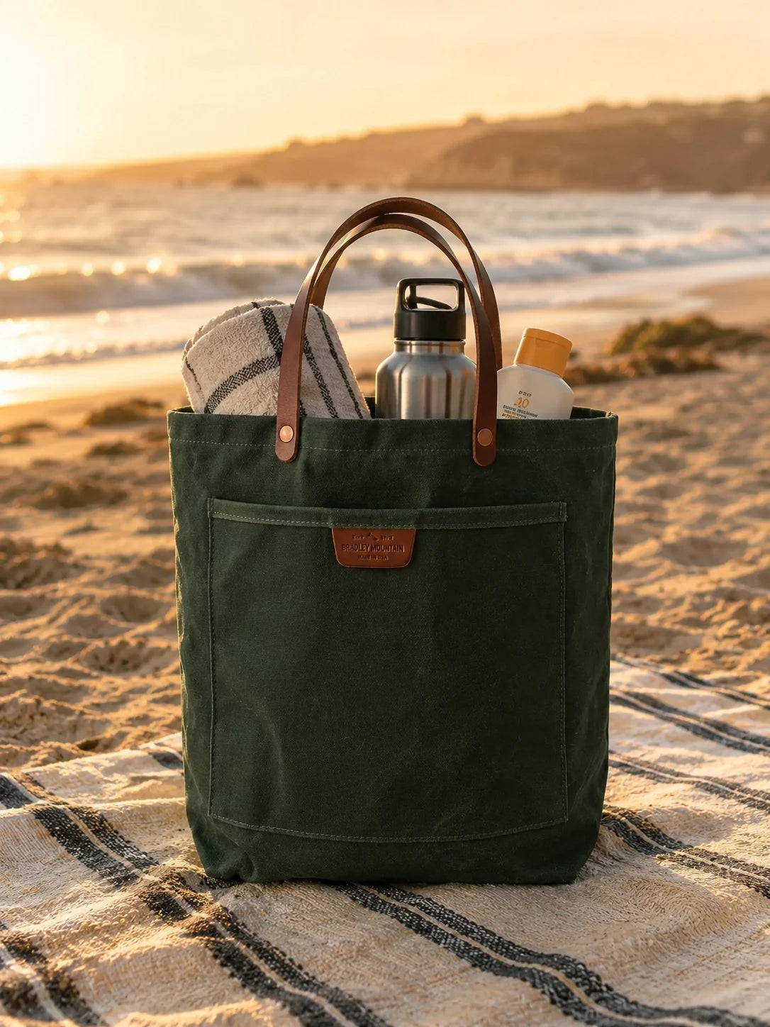 Coal Tote on a striped blanket at the beach with a towel, bottle and sunscreen.